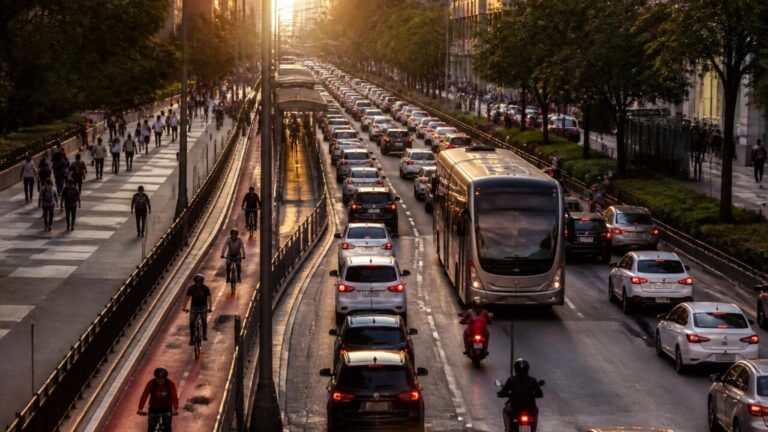 Rua urbana com tráfego misto em horário de pico, incluindo carros, ônibus e ciclistas.